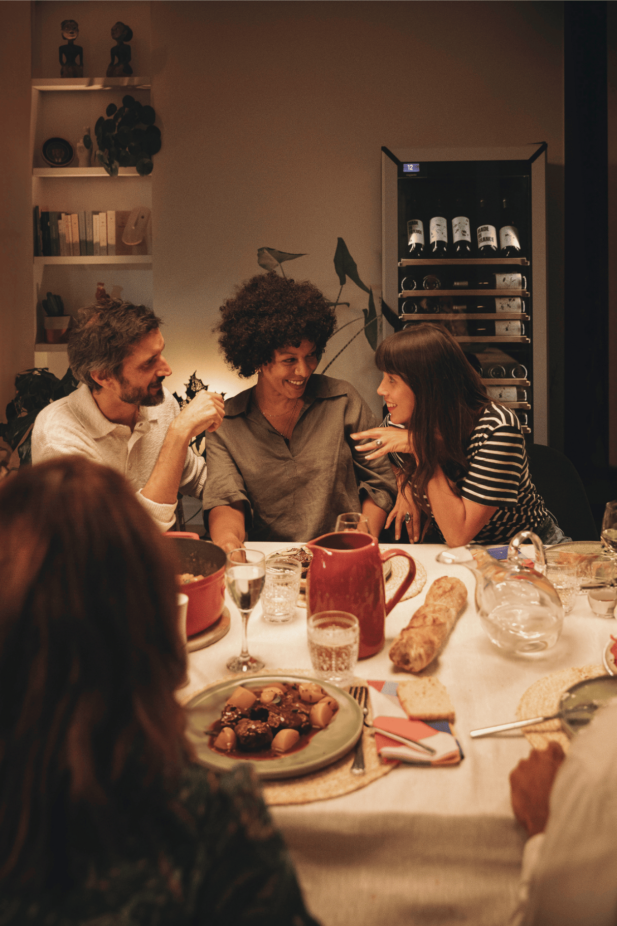 Friends enjoying a dinner with food on the table and a goguette wine frige in color white, large unit with presentation row in the background.