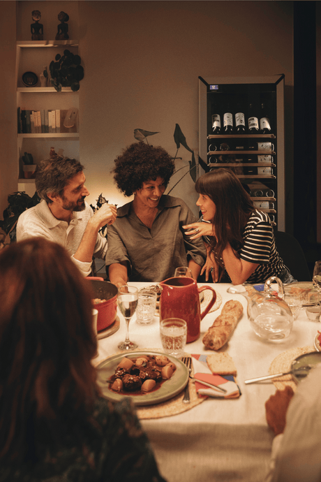 Friends enjoying a dinner with food on the table and a goguette wine frige in color white, large unit with presentation row in the background.